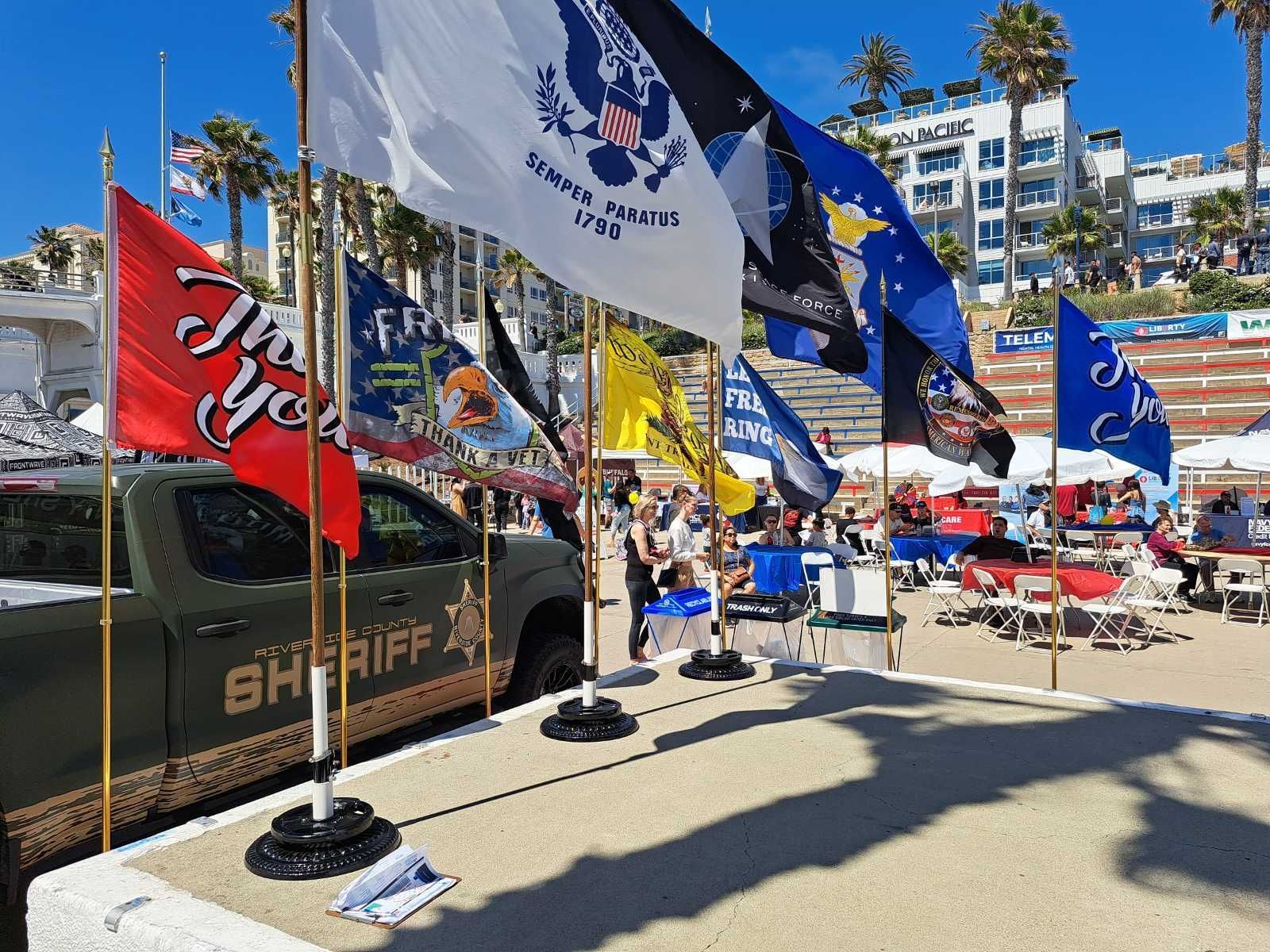 Flags and a Sheriff's vehicle on the beach, people seated at tables, sunny day.