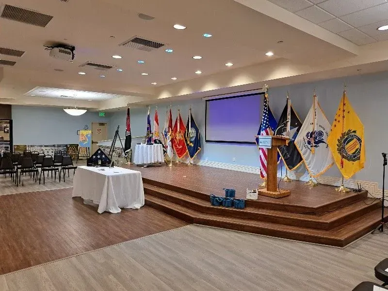 Room set up for ceremony, stage with flags and podium, empty chairs, and memorial table.