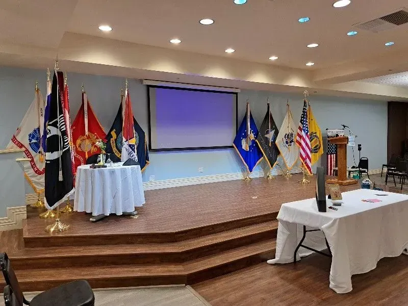 Stage with flags and a screen, set for an event; a small table is on the stage left and another is on the stage right.