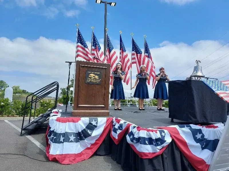 Four women singing on a stage decorated with American flags. Podium, bell, and patriotic bunting. Sunny, outdoor setting.