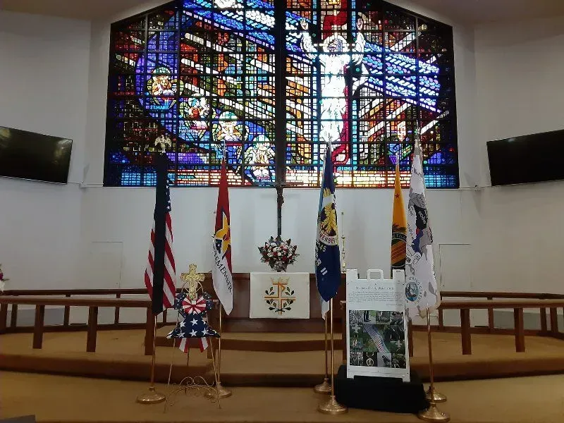 Altar with flags, stained glass window, cross, and floral arrangements inside a church.