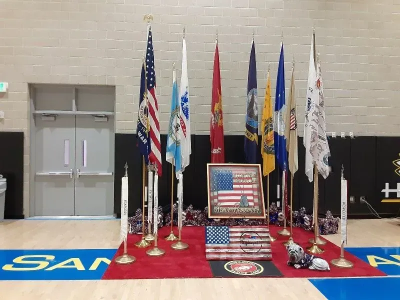 Flags of the US military branches displayed on a red carpet in a gymnasium, with patriotic plaque.