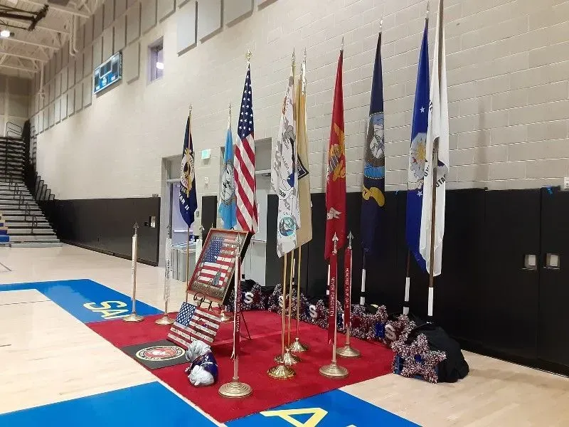 Flags of different military branches and the United States on display on a red carpet in a gymnasium.