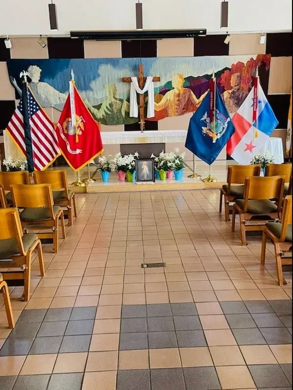 Interior of a chapel with flags, flowers, and a crucifix on a raised platform. Wooden chairs are in rows.