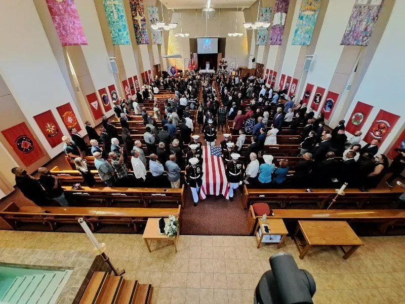 Funeral service with a flag-draped casket carried through a church; attendees standing in rows of pews.