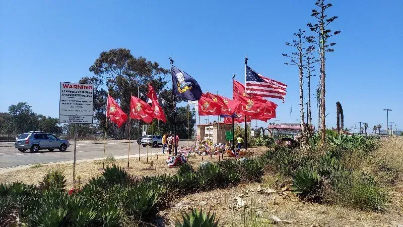 Flags and a memorial on a roadside with cars and a sign under a blue sky.