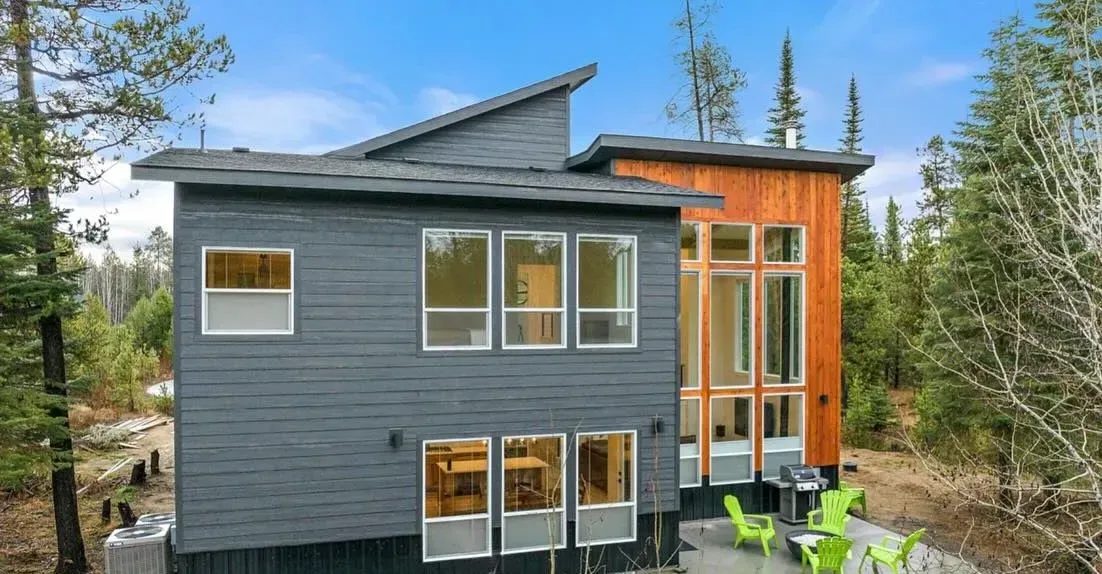 Modern house exterior with gray siding, large windows, and a wood-paneled section. Green chairs sit on a patio.