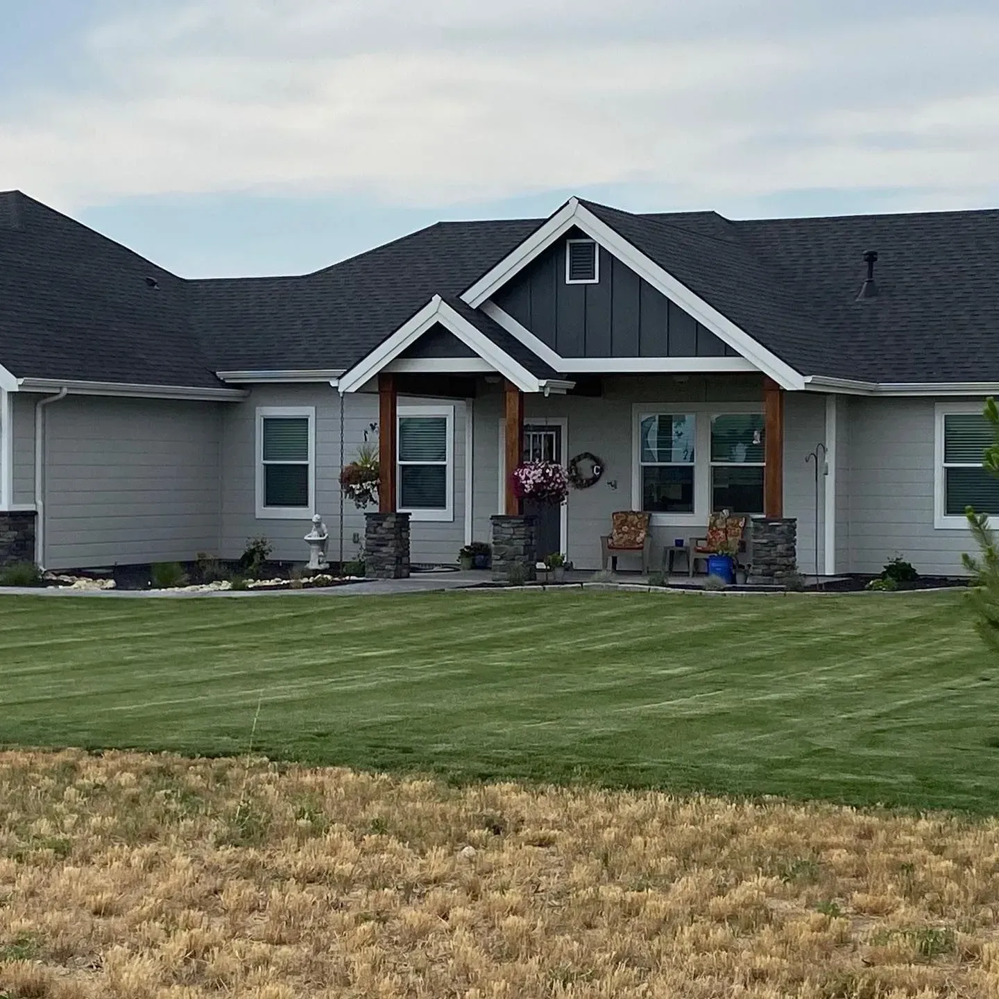 Gray house with dark roof and green lawn. Brown porch columns, small flower pots, and a wreath.