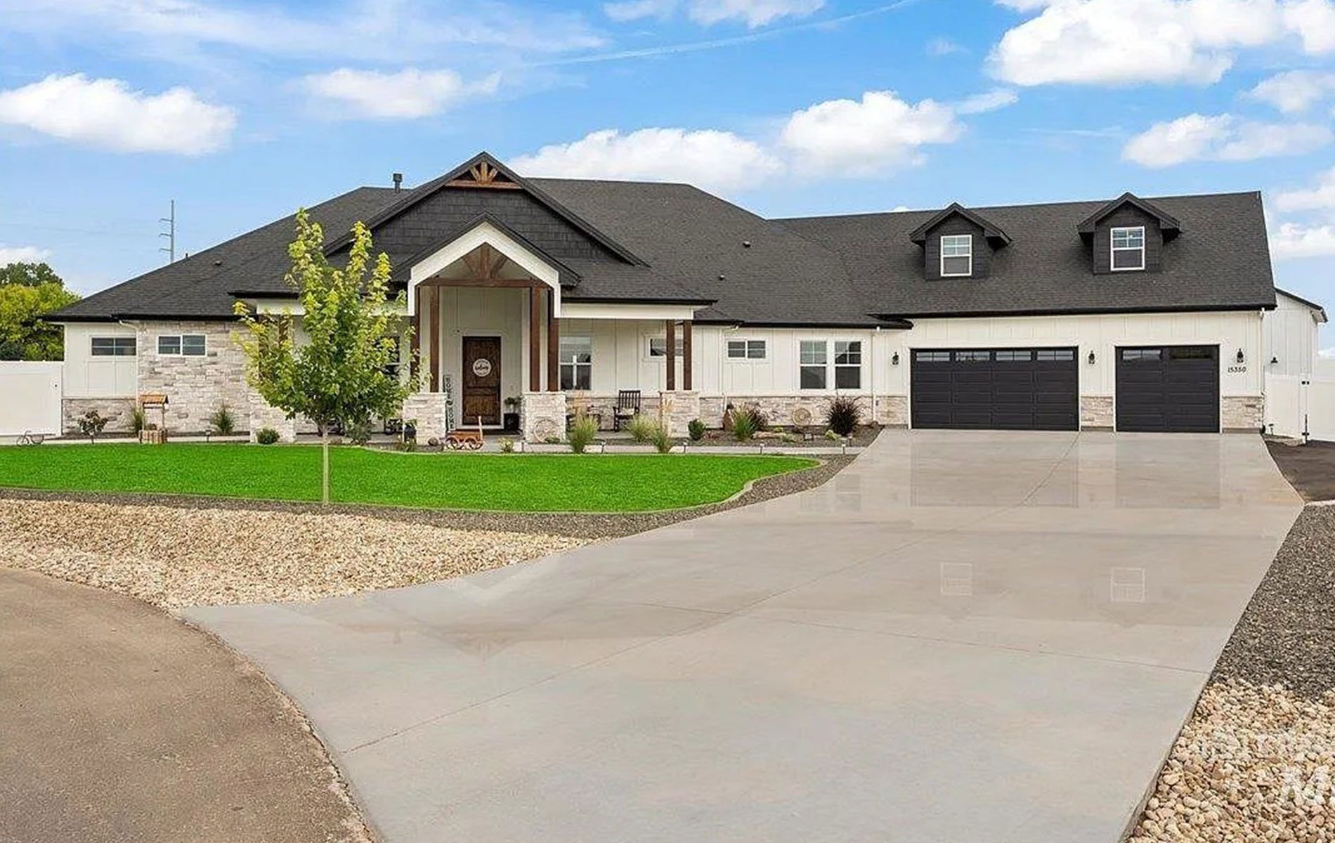 White house with black garage doors, gray driveway, and manicured lawn under a blue sky.