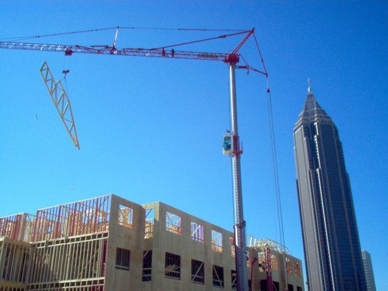 Construction crane lifting materials, wooden building frame, skyscraper in background against a clear blue sky.