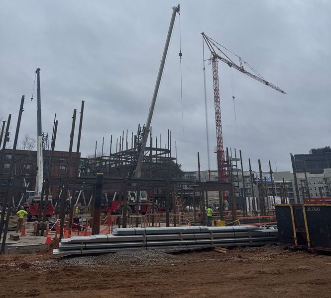 Construction site with steel framework and two cranes. Workers in hard hats. Cloudy sky.