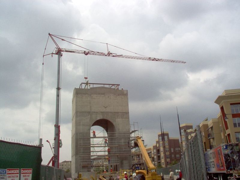 Construction site with crane over a concrete archway, workers present. Cloudy sky, other buildings in background.