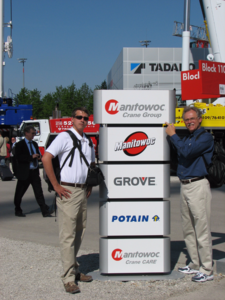 Two men posing near a stack of Manitowoc Crane Group signs at an outdoor event.