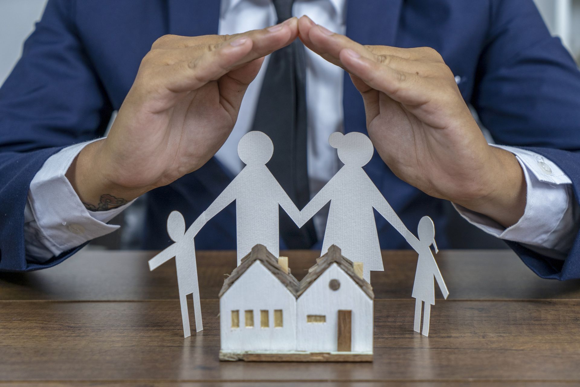 Hands shielding paper family and house model on a table, symbolizing home protection.