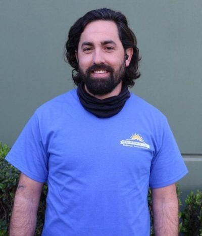 Smiling person in a blue T-shirt and dark neck gaiter standing against a gray wall