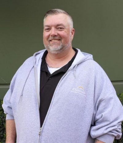 Smiling man in a light gray hoodie standing against a green wall