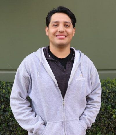Person smiling in a gray hoodie, standing outdoors against a green wall and shrubs.
