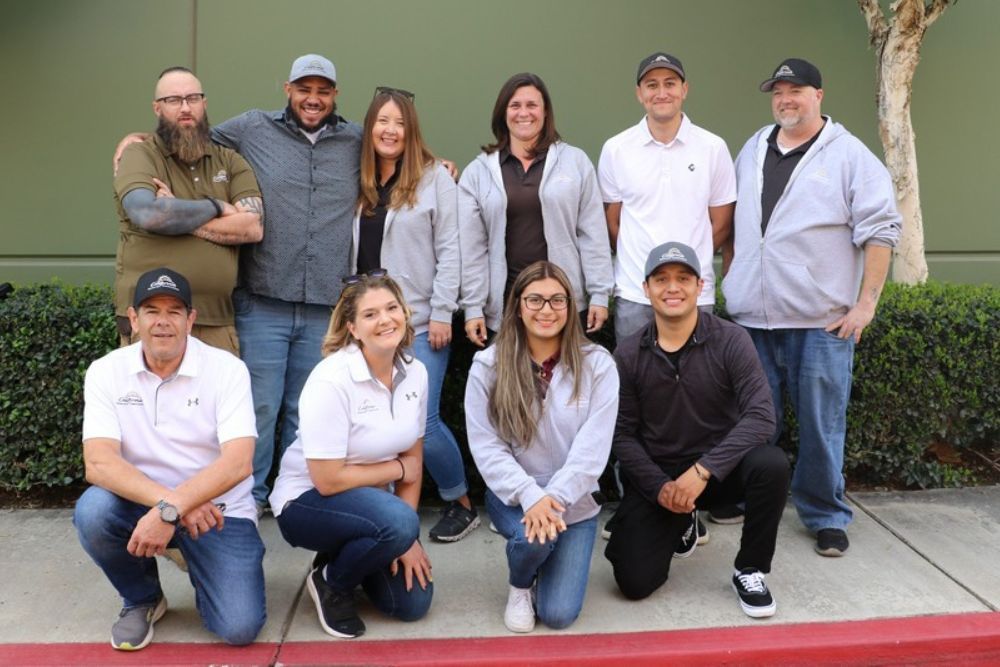 Group of nine coworkers posing outdoors in front of a building, smiling at the camera.