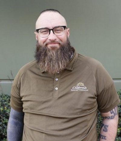 Smiling bearded man in glasses wearing an olive polo shirt against a plain wall