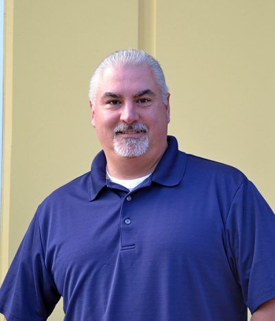 Smiling man in a blue polo shirt standing against a light yellow wall.