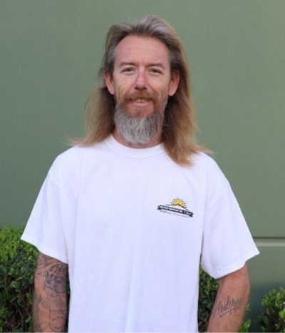 Person with long hair and beard wearing a white T-shirt, standing outdoors against a green wall.