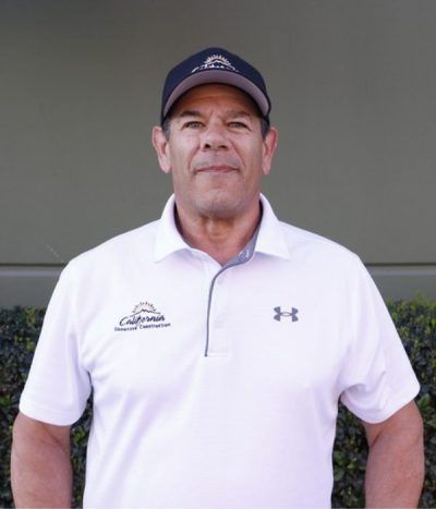 Man standing against a gray wall, wearing a navy cap and white polo shirt with logos
