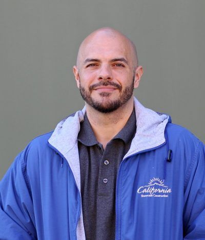 Man in a blue jacket over a gray polo, standing against a plain gray background, facing the camera.