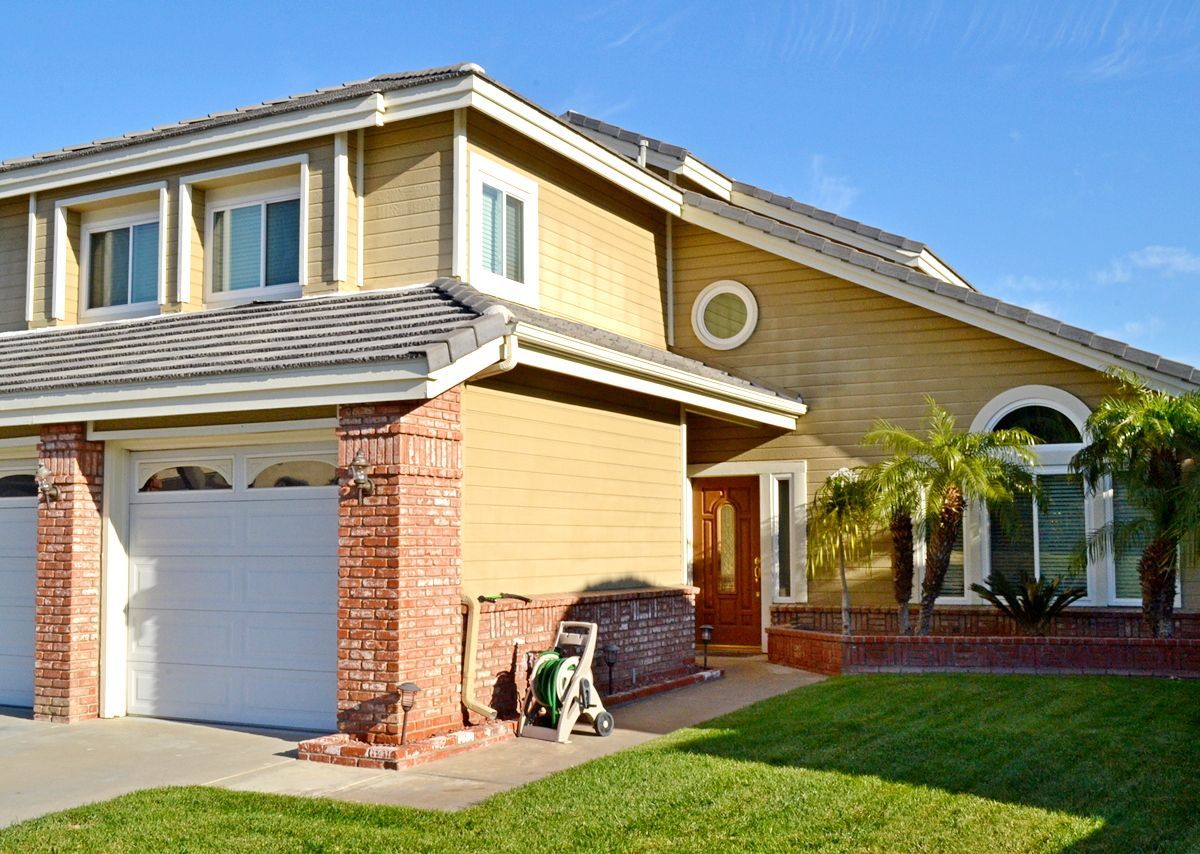 Suburban two-story house with beige siding, brick accents, garage, and a palm tree in the front yard
