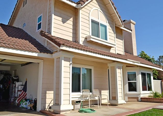 Suburban tan house with brown roof, front patio chairs, and open garage under a sunny sky