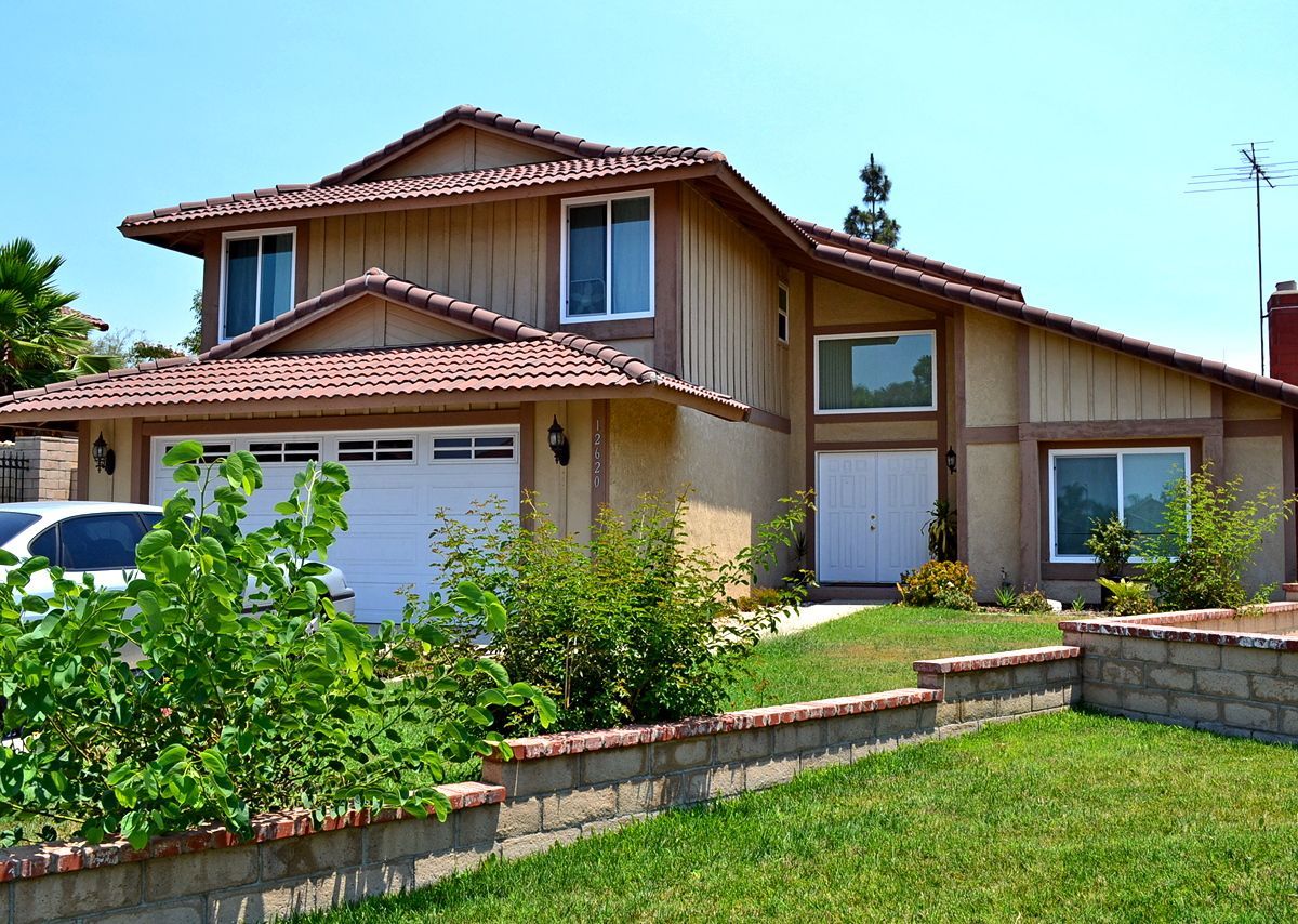 Two-story beige house with red tile roof, garage, and front garden on a sunny day