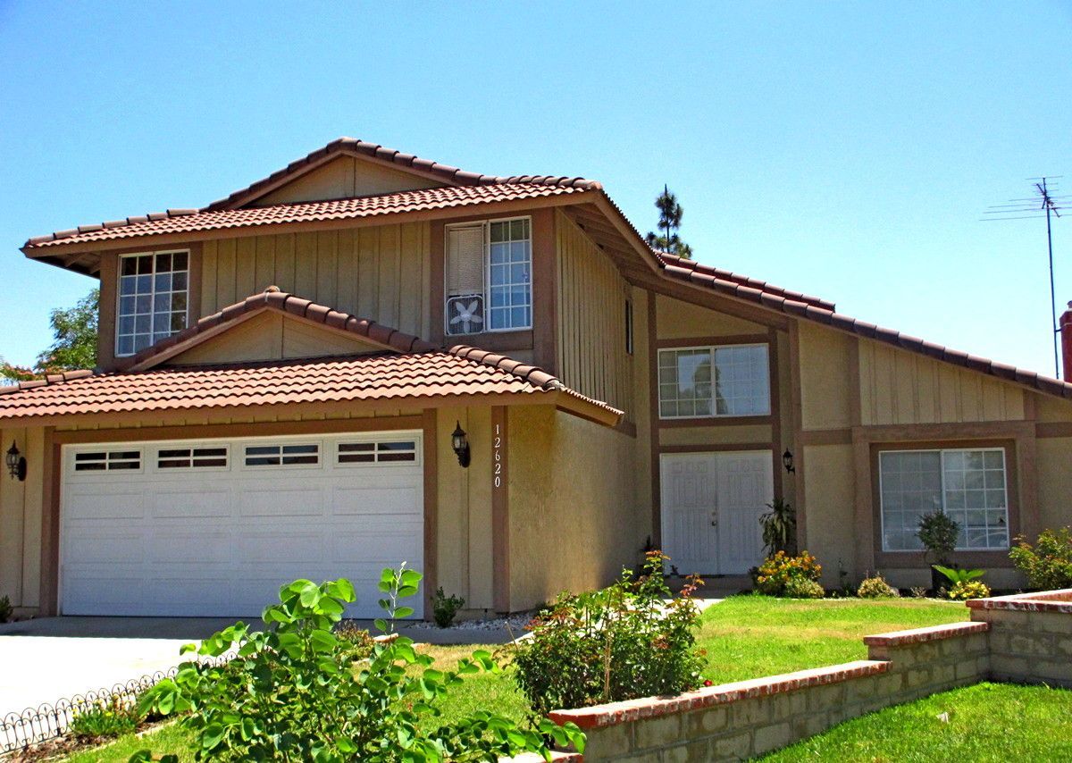 Two-story beige suburban house with a white garage, red-tiled roof, and green lawn