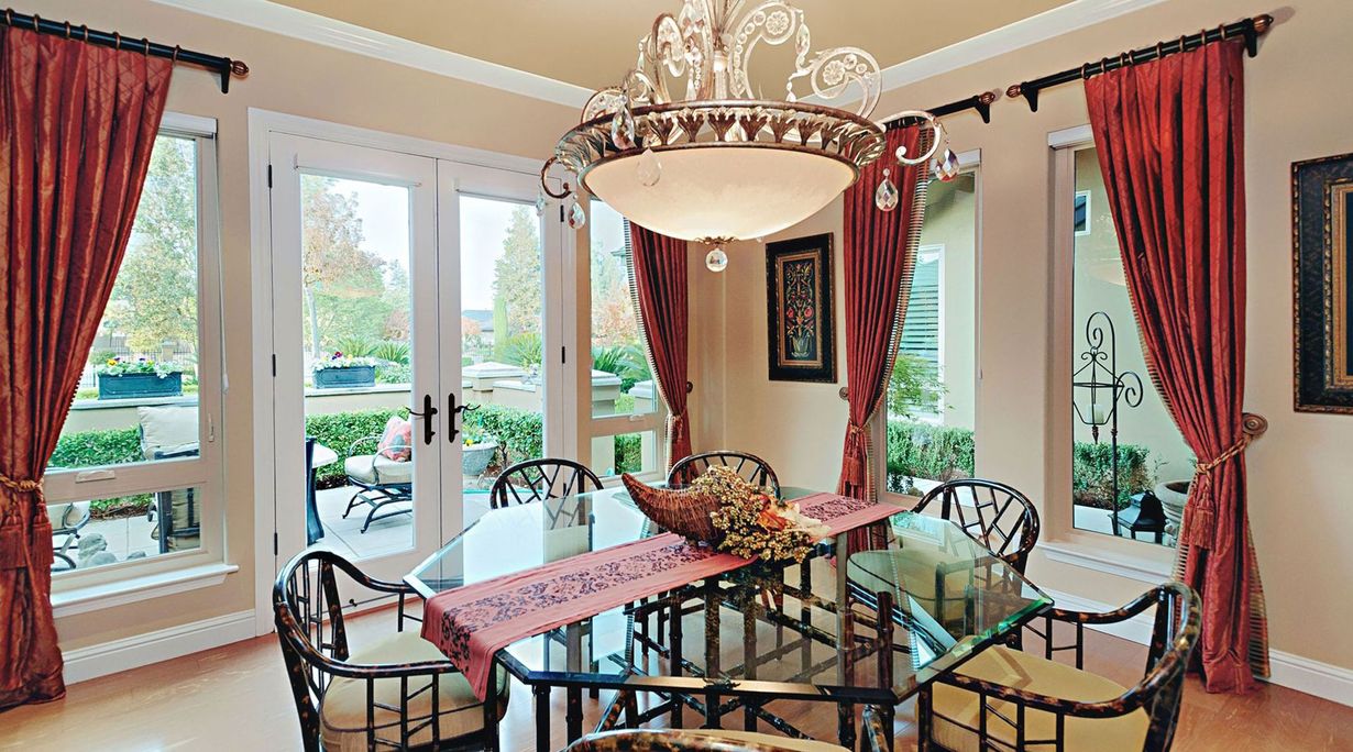 Elegant dining room with glass table, ornate chandelier, red curtains, and large windows overlooking greenery