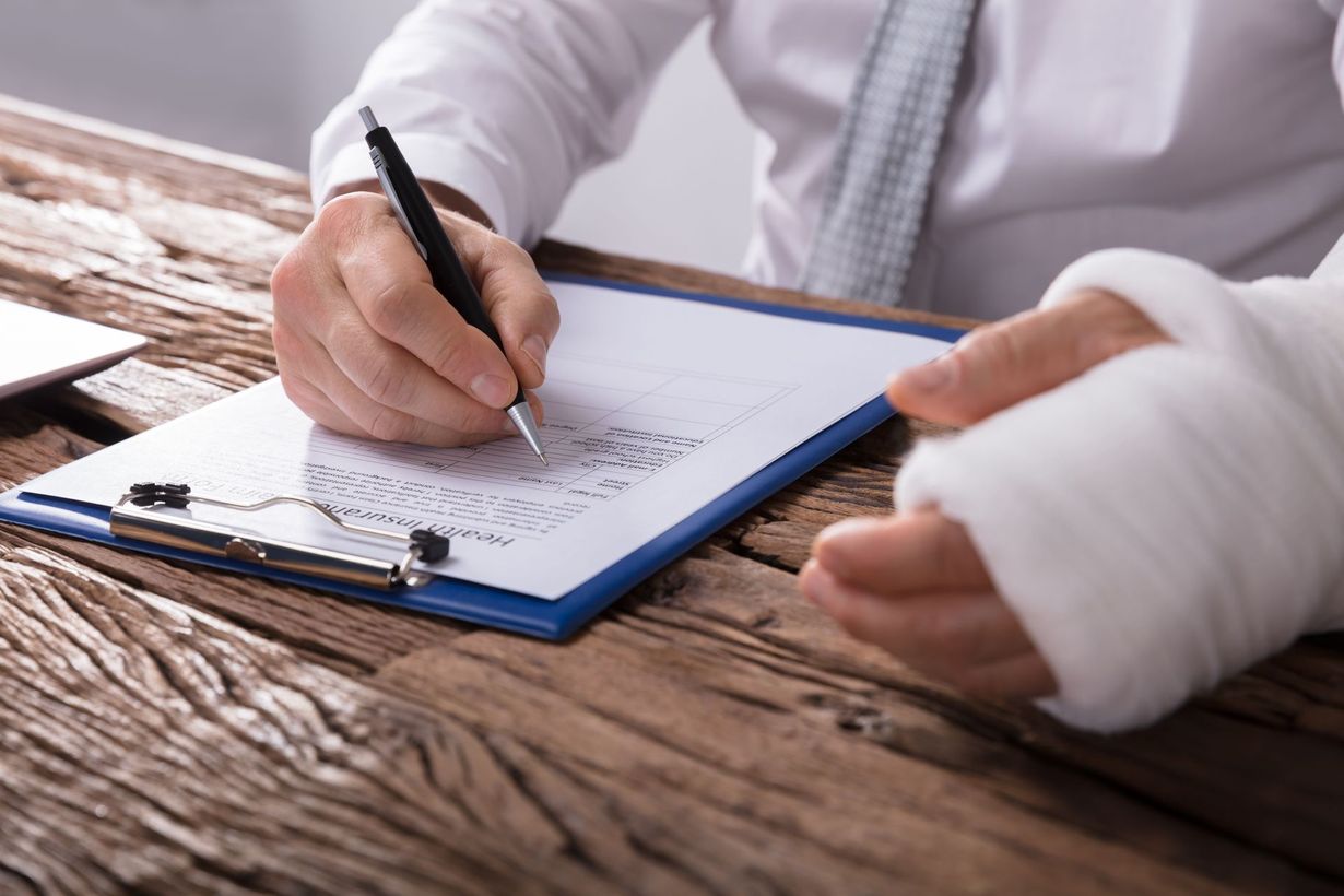 A person in business attire with their left arm in a white cast writes on a document attached to a blue clipboard.