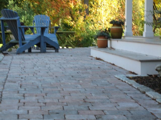 A brick walkway leading to a porch with blue chairs and potted plants