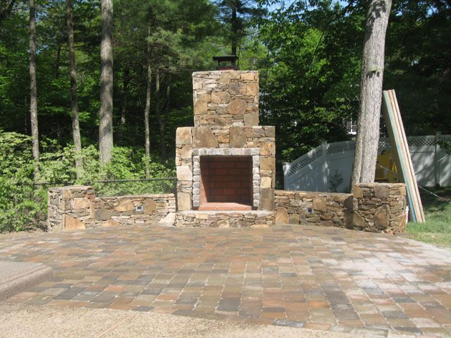 A stone fireplace is sitting on top of a brick patio surrounded by trees.