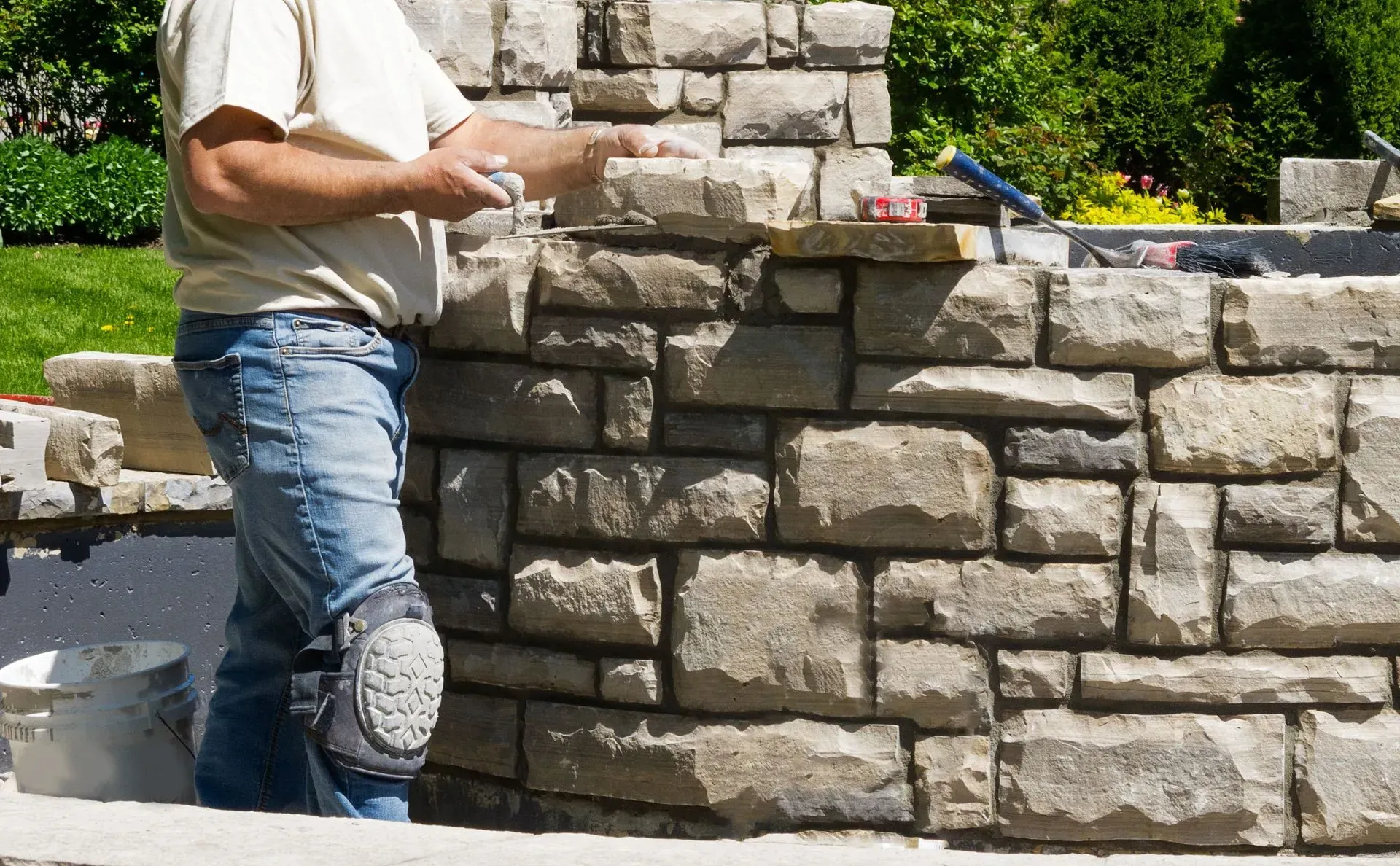 A person building a stone wall, wearing jeans and knee pads, outdoors.