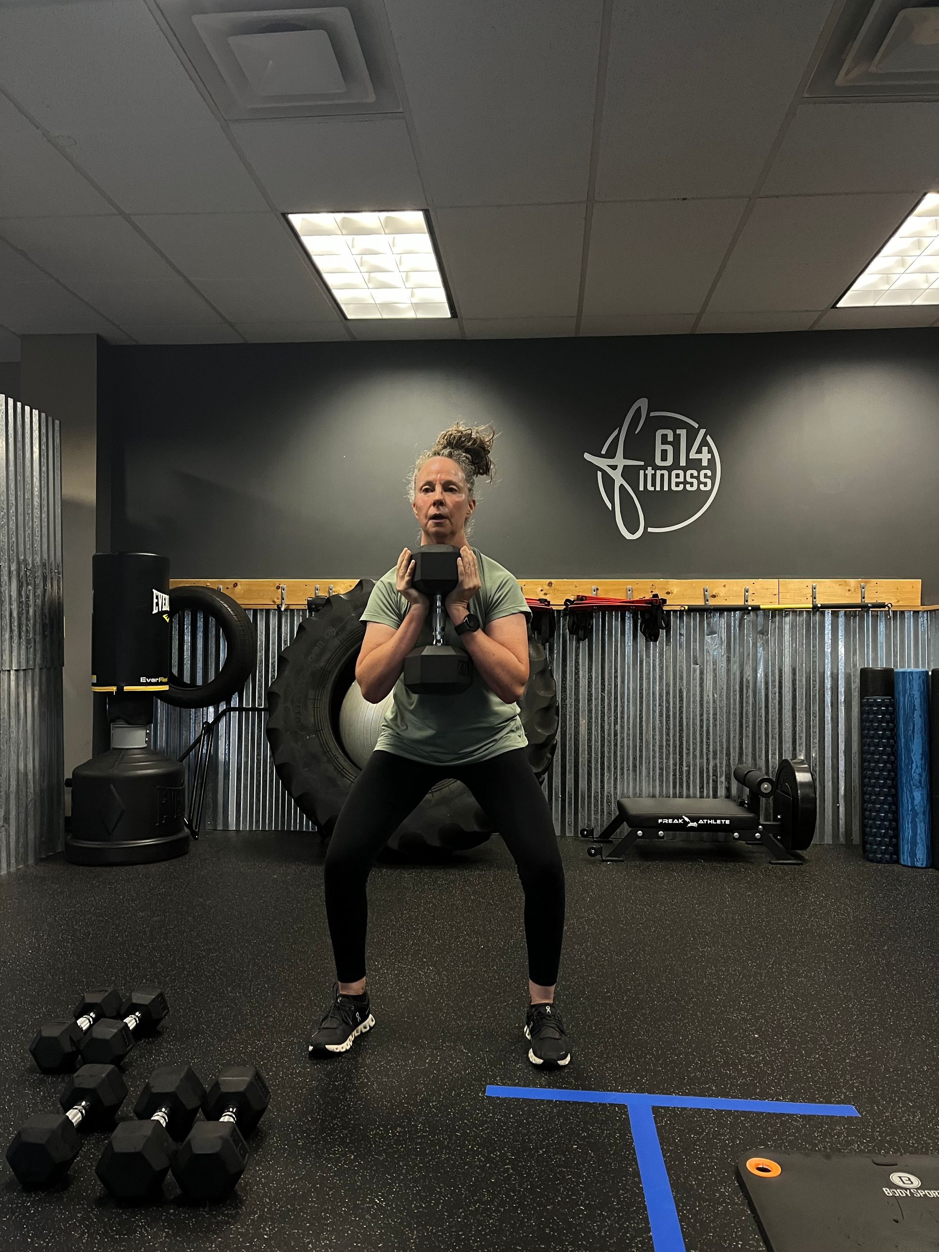 Woman in a gym performing a dumbbell squat, holding the weight near her chest, in front of a tire and weights.