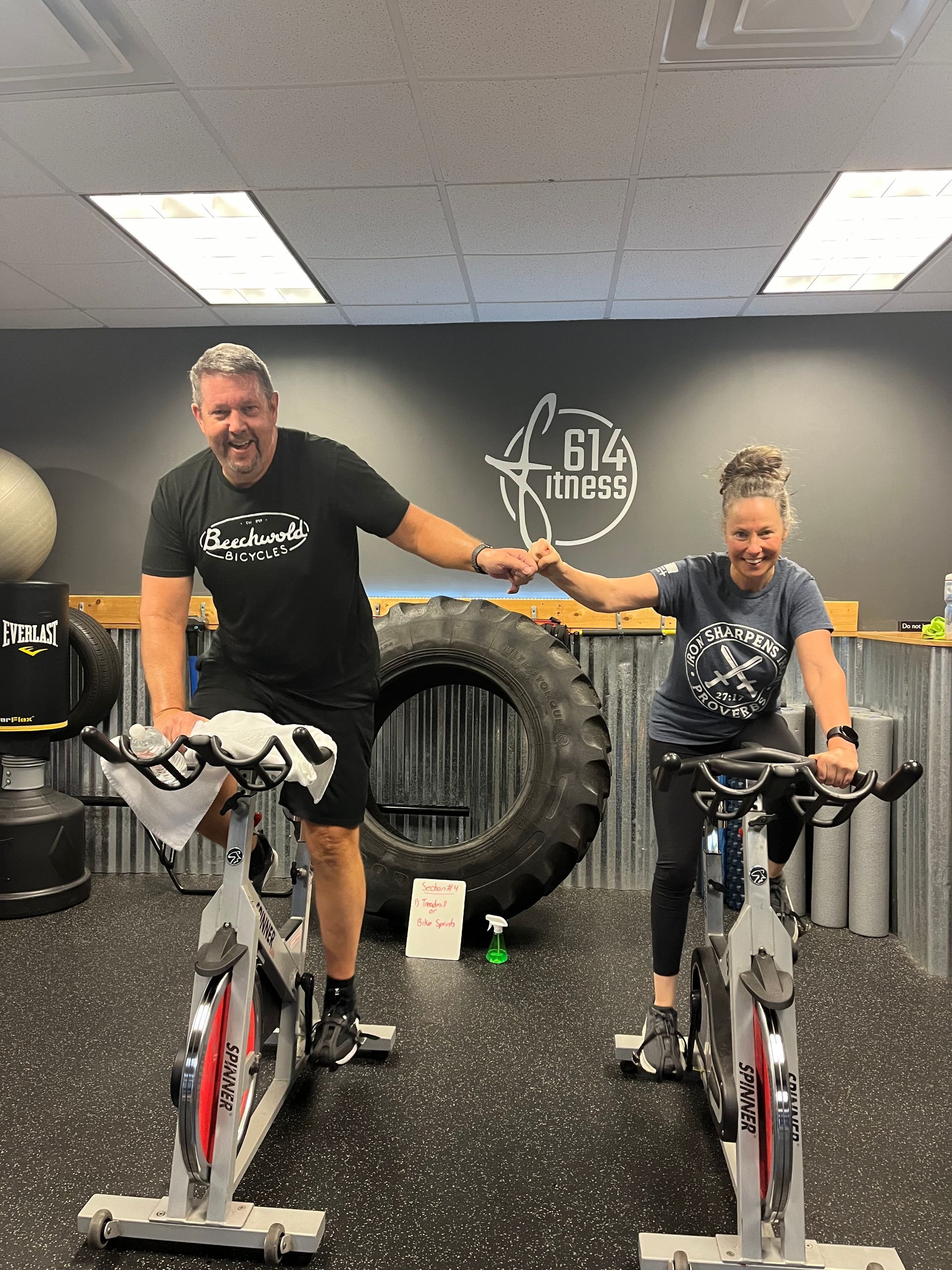 Two people fist-bumping while riding stationary bikes in a gym. Dark gray walls, tire, and 614 Fitness logo visible.