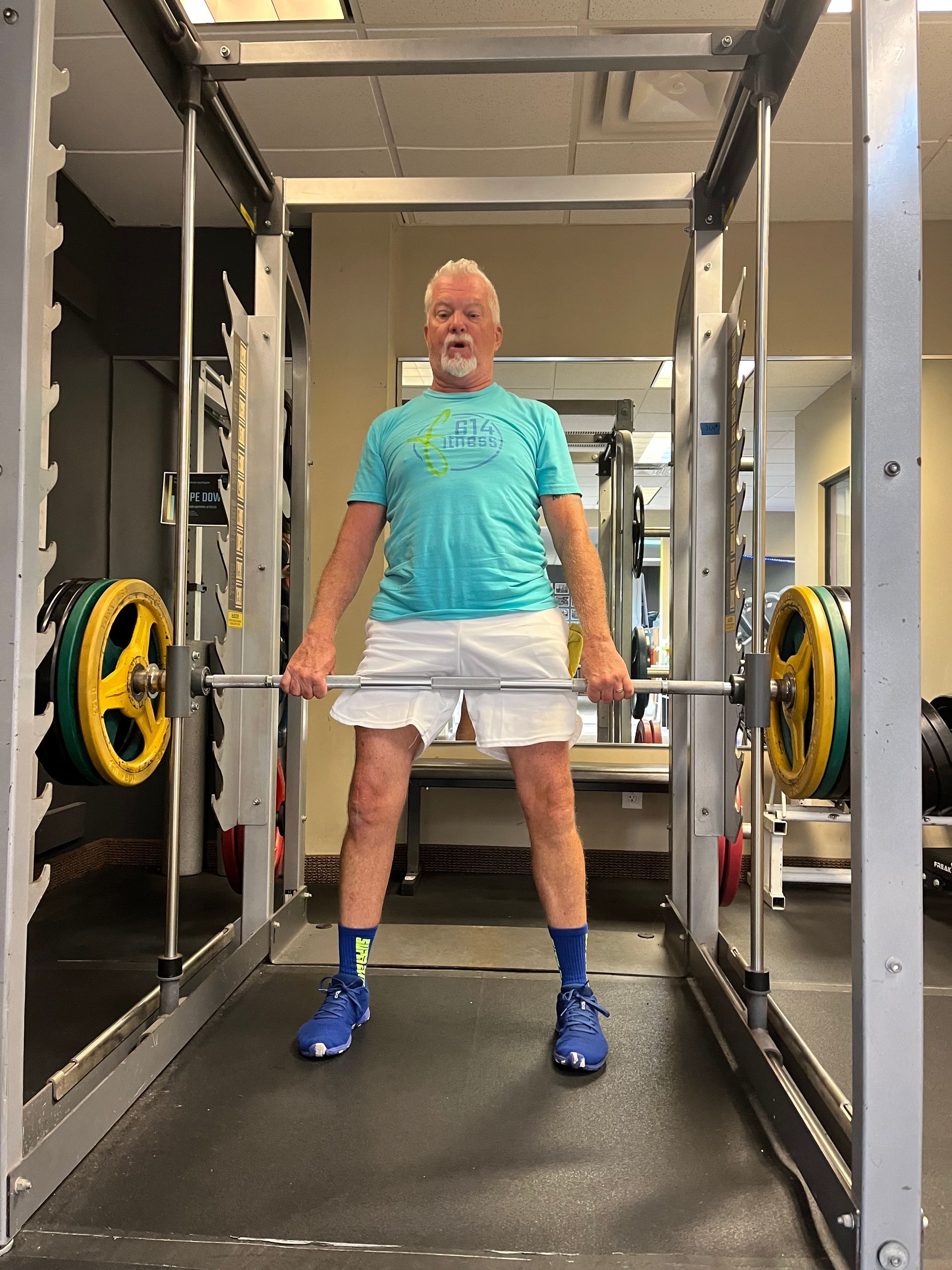 Man in gym holding barbell, standing in Smith machine. He wears shorts, t-shirt, and blue sneakers.