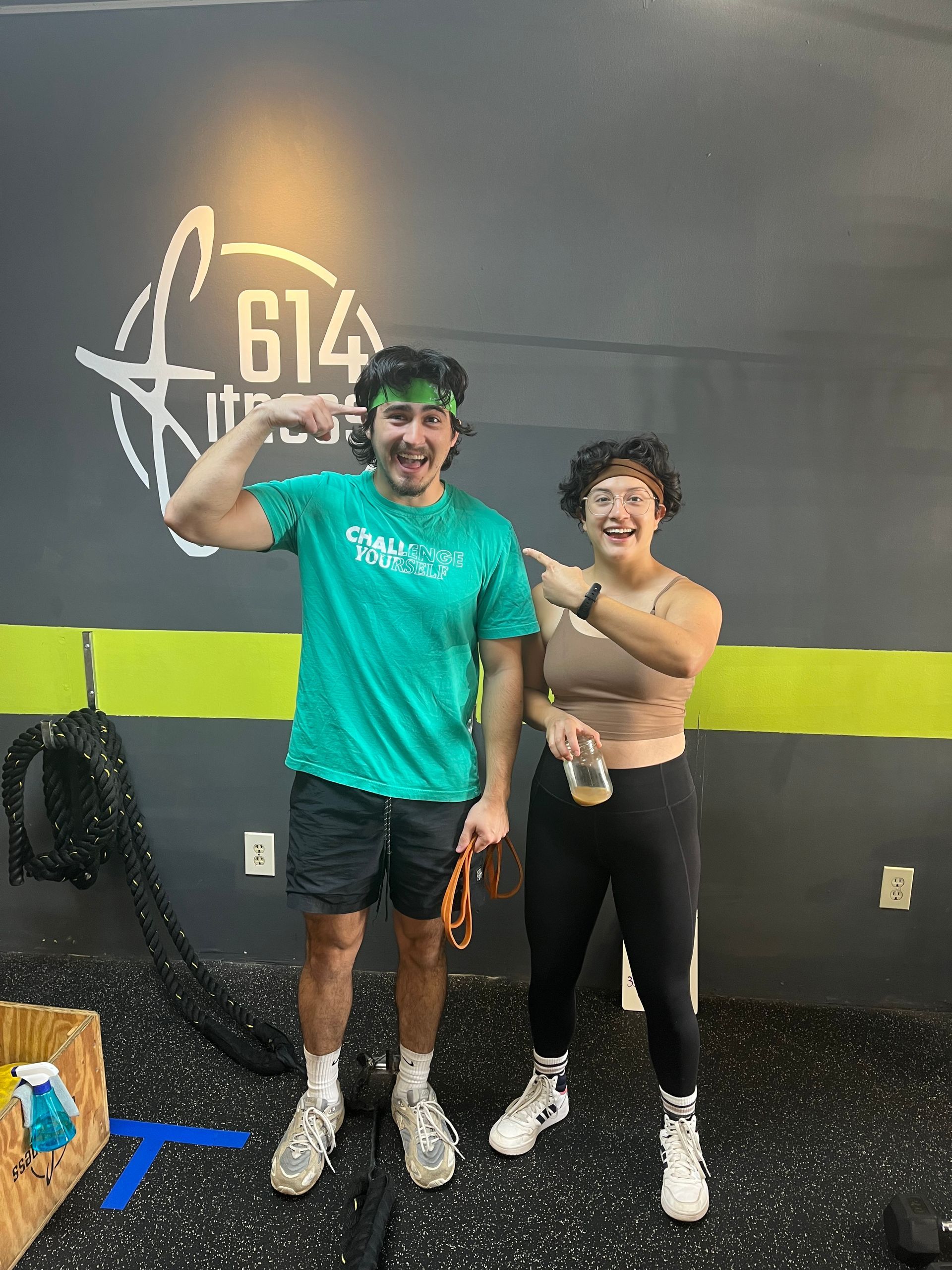 Two people smiling at a gym. Man flexes, woman points. Green and black clothing. 