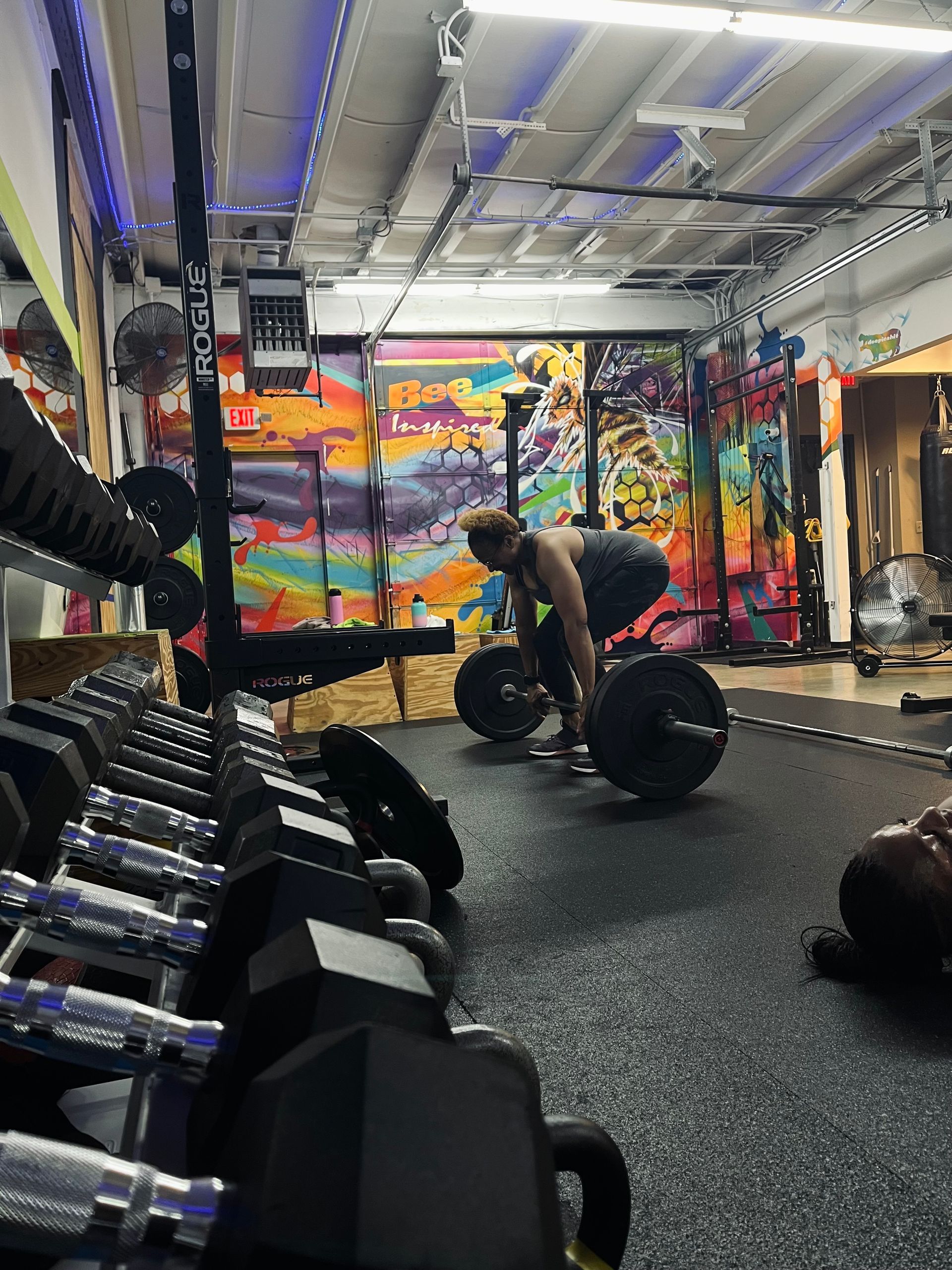 Person lifting a barbell in a gym, focusing on the deadlift. Weights and equipment are in the foreground. Colorful graffiti art in the background.