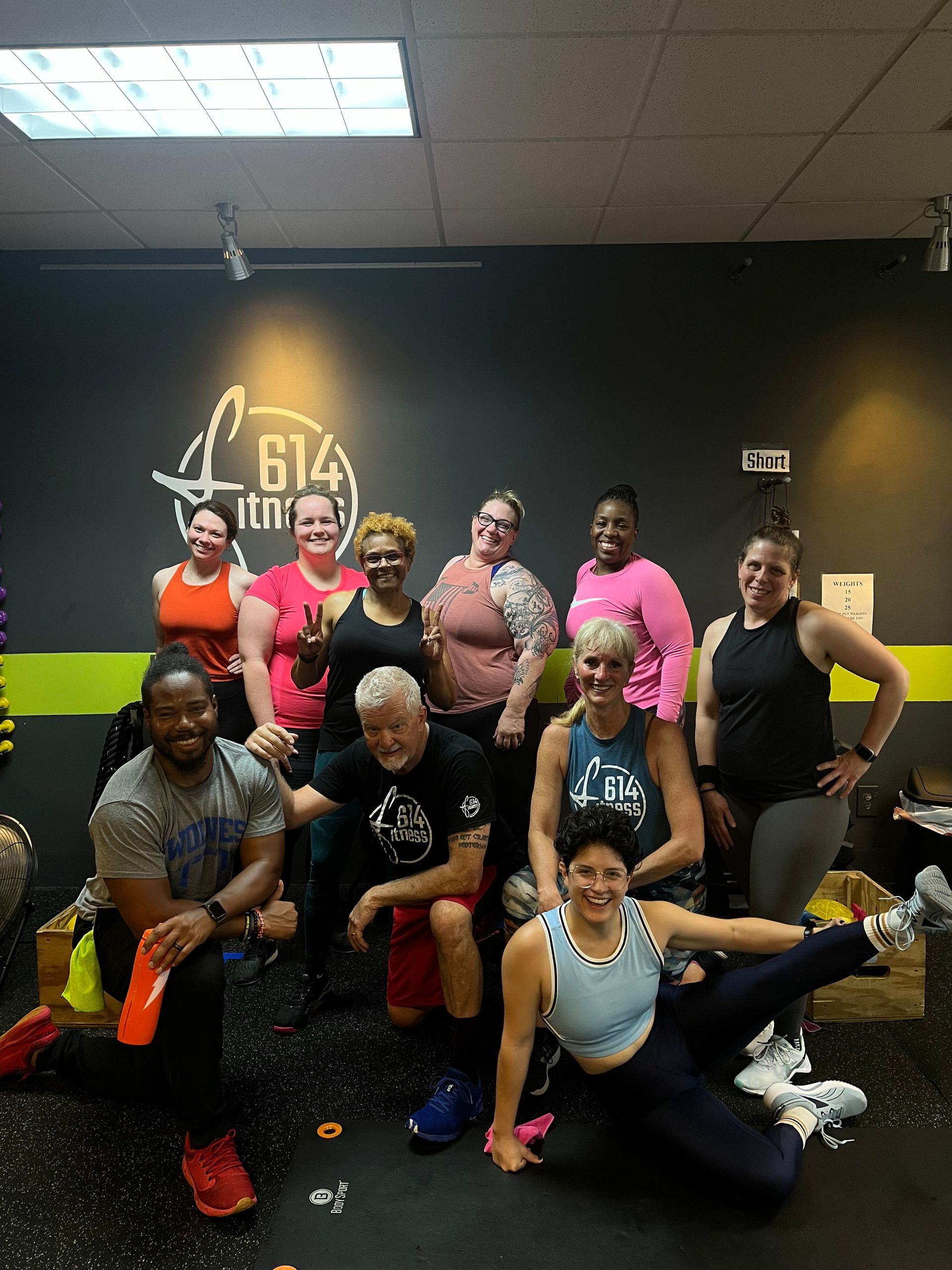 Group of people smiling, posing in a fitness studio. The background features a logo and black and lime green walls.