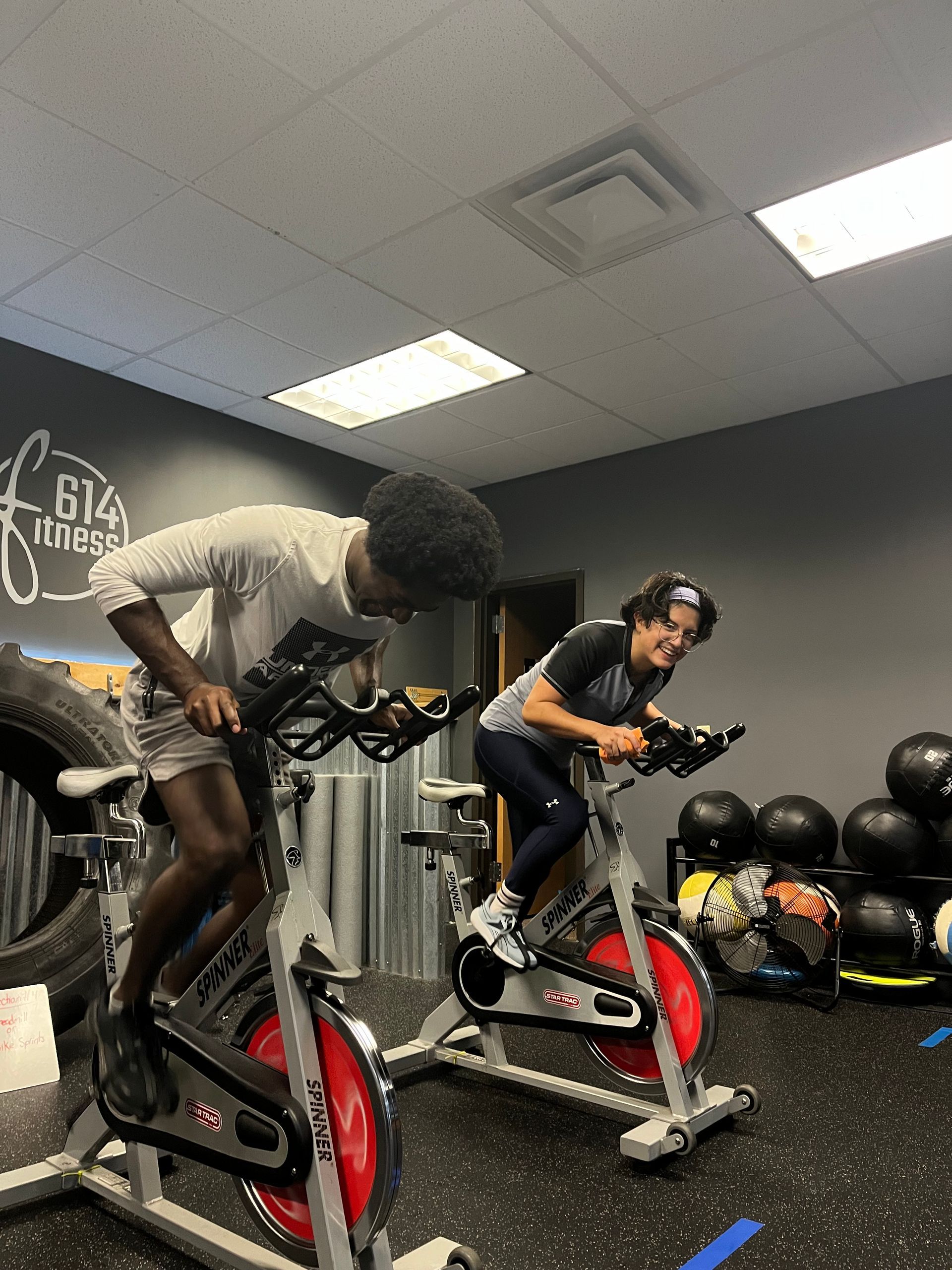 Two people riding stationary bikes in a gym. Dark-haired individual in gray shirt, other in black, both pedaling.