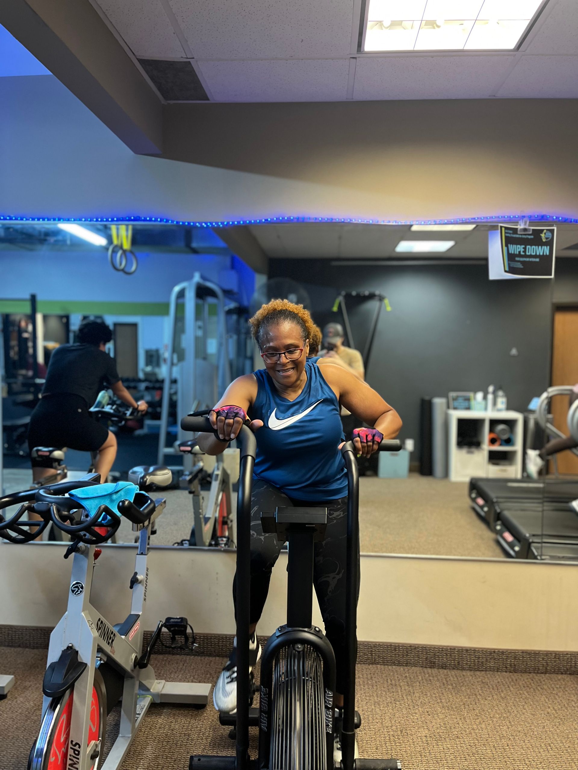 Woman in blue shirt using an elliptical machine at a gym. A person cycles in the background.