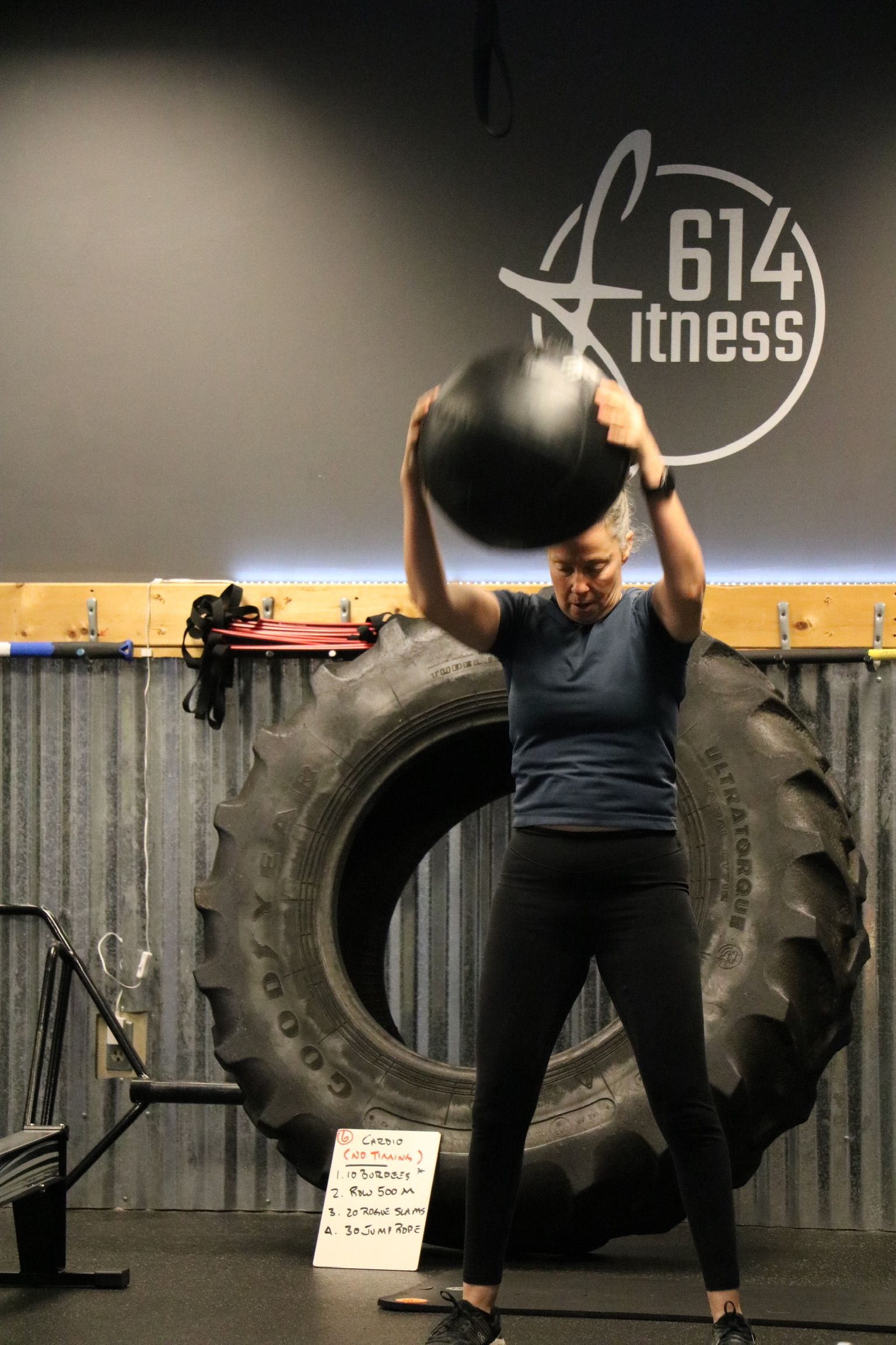 Woman in gym, overhead ball slam. Black ball, tire background, 