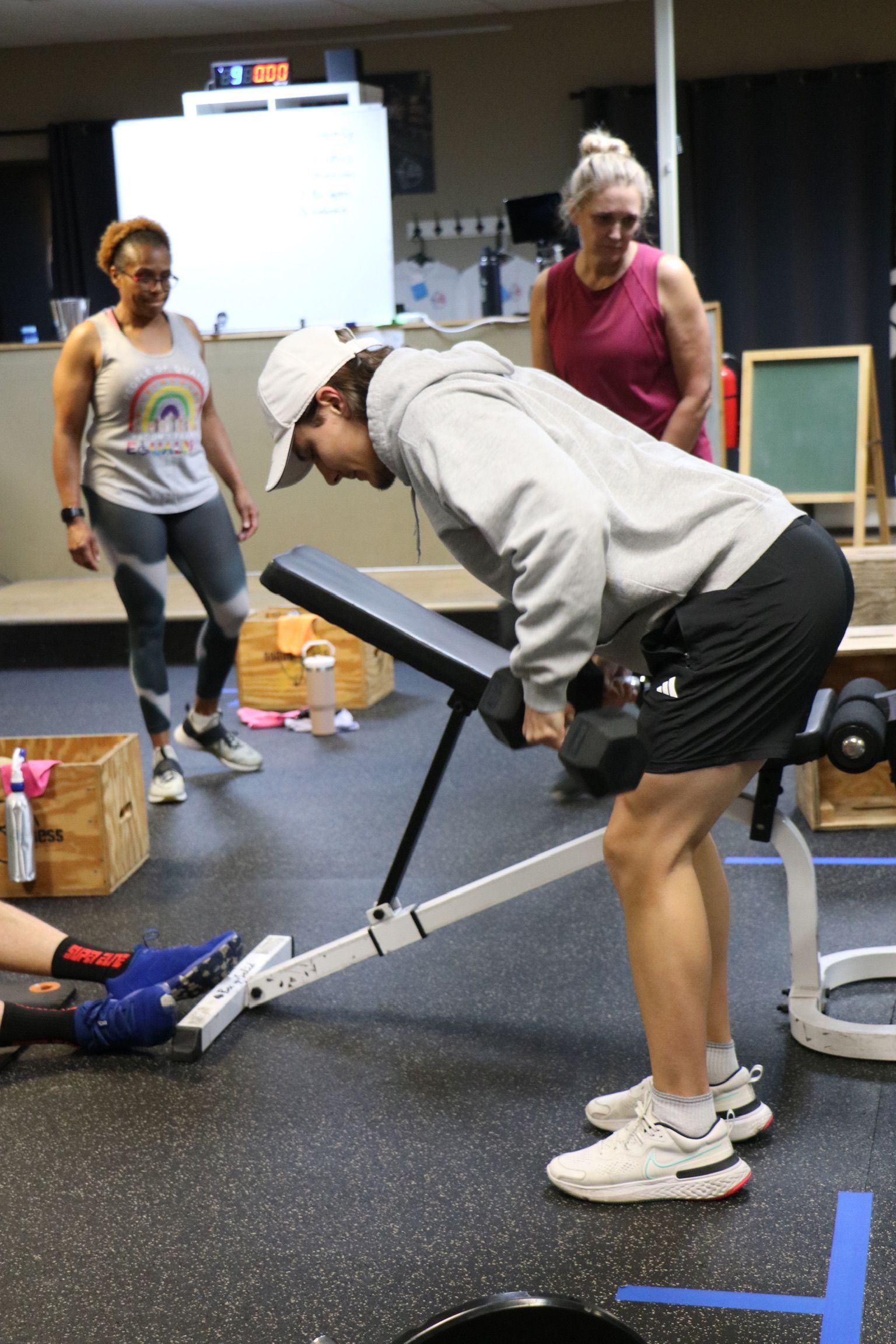 Man doing dumbbell rows on a bench in a gym. Two women watch.