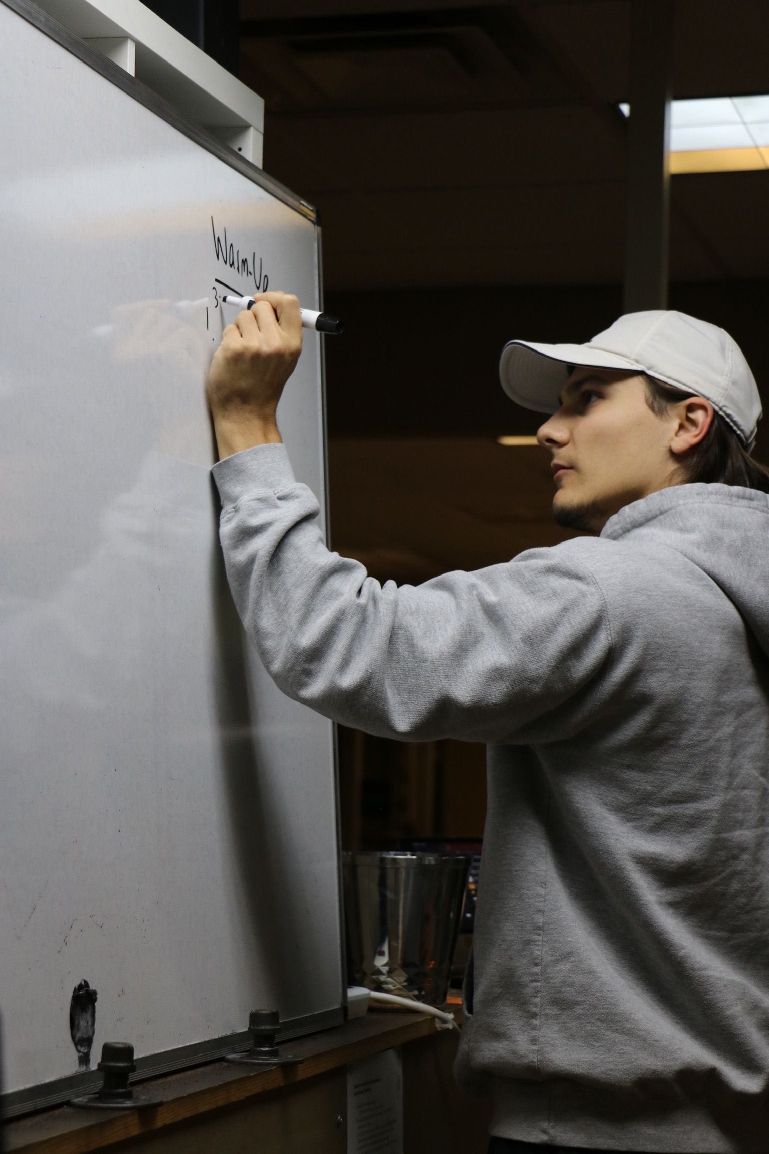 Person in a gray hoodie and cap writes on a whiteboard with a marker indoors.