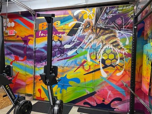 Colorful graffiti art of a bee on a garage door inside a gym, with a rowing machine in the foreground.