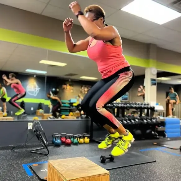 Woman in athletic wear jumps onto a wooden box in a gym, neon green sneakers, other people visible in background.