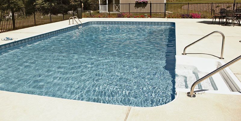 Rectangular swimming pool with steps and metal handrails; concrete deck, fenced backyard.